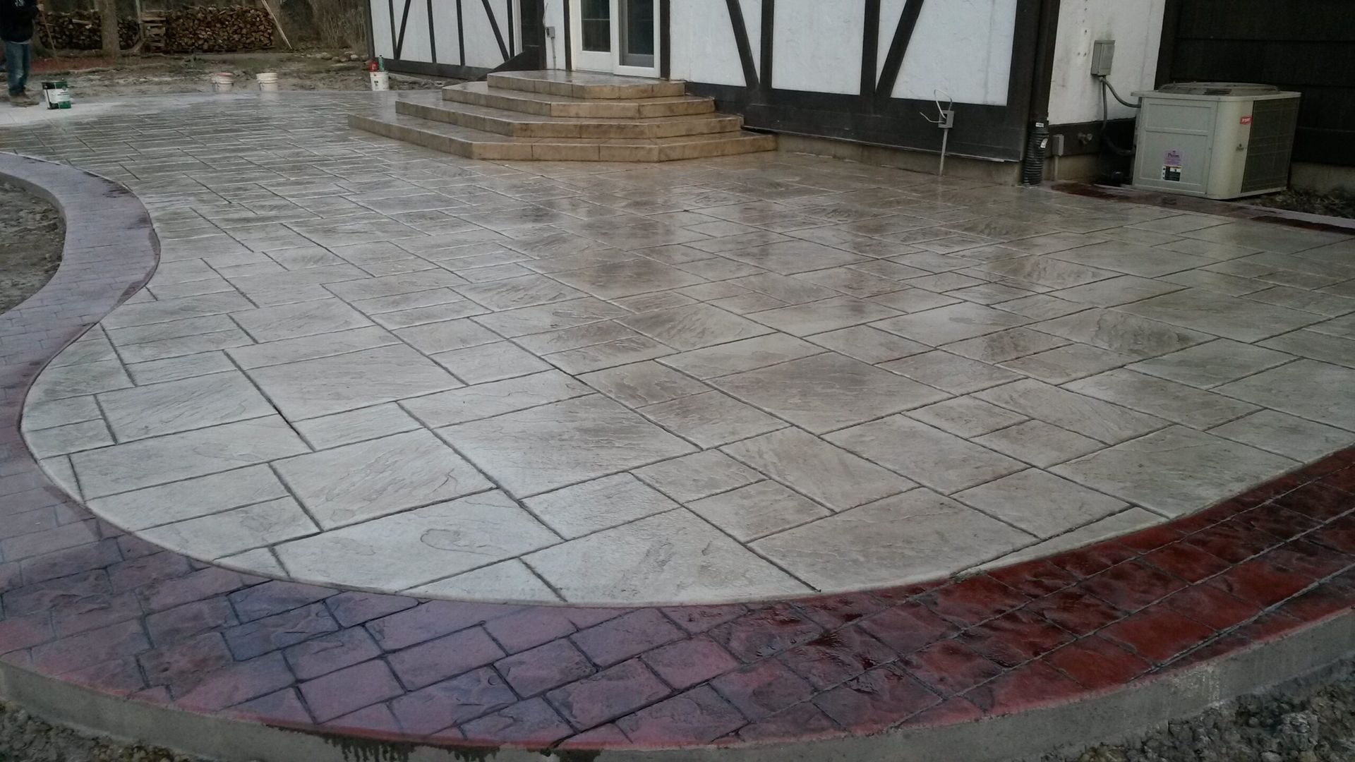 Concrete patio with brick-red border and a large main area, wet from rain, in front of a house.