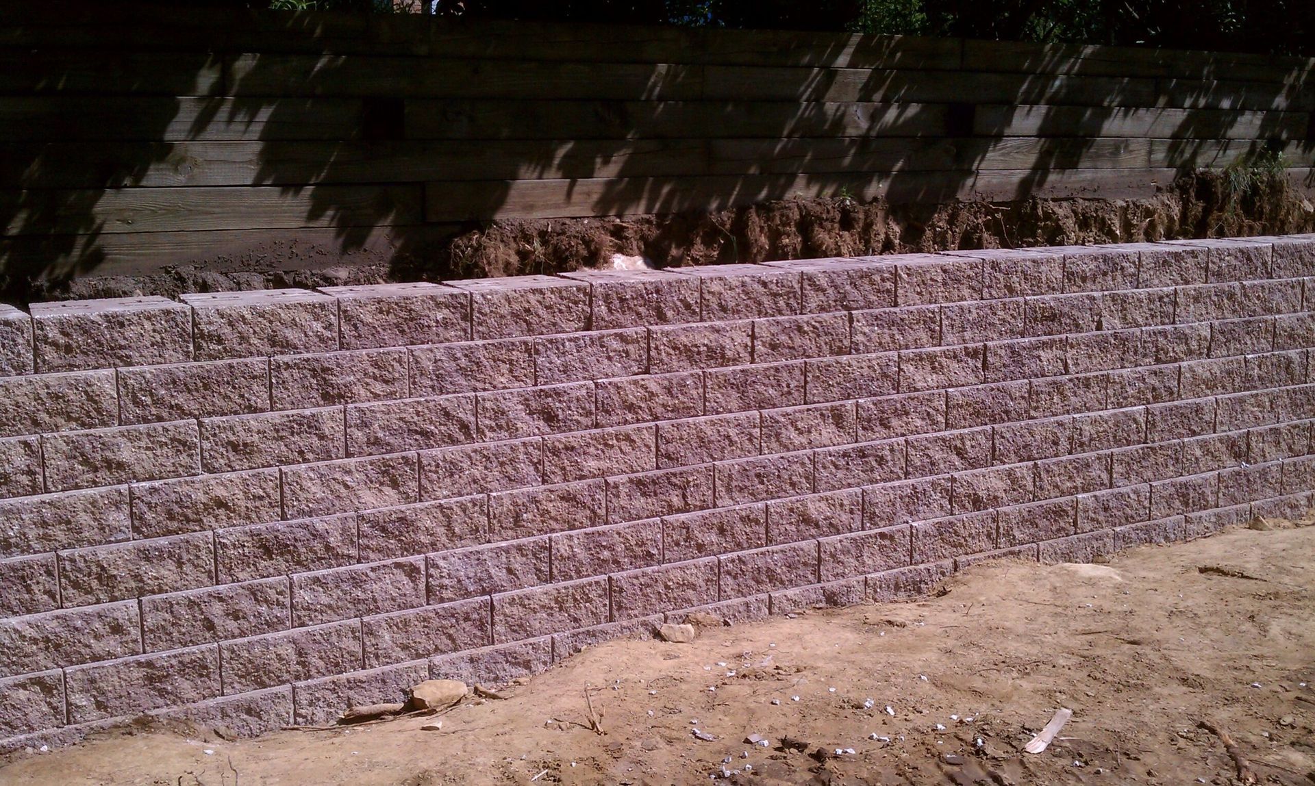 Retaining wall made of reddish-brown textured blocks, set against a dark wooden fence and dirt.