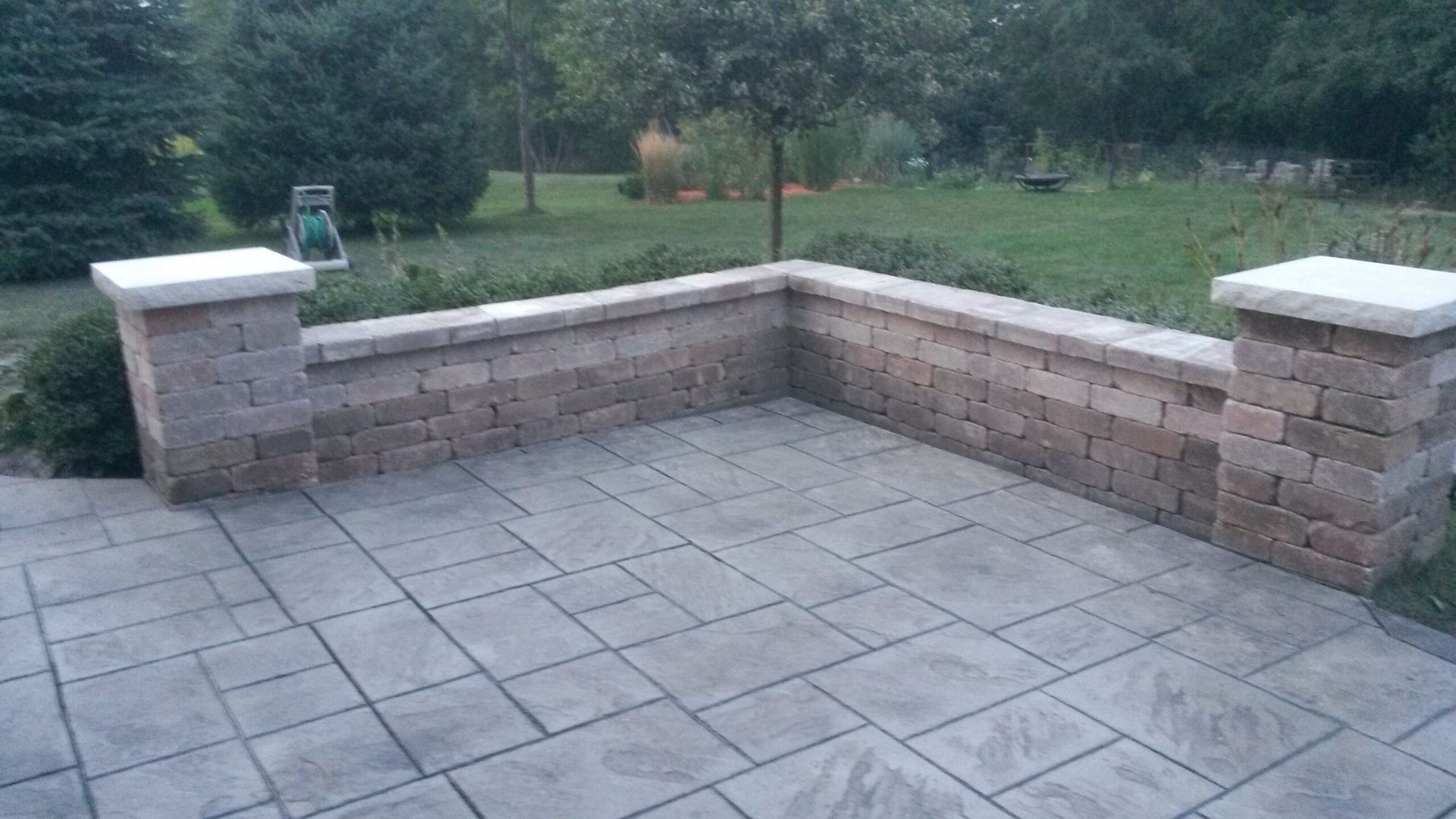 Stone patio with low brick wall and square pavers. Green yard and trees in the background.