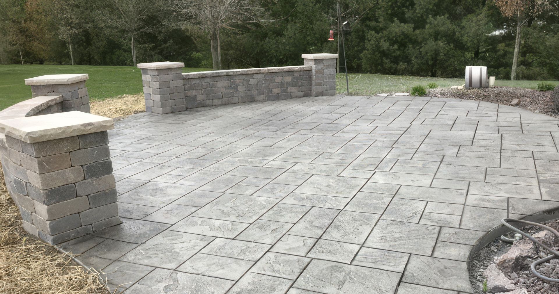 Stone patio with low brick walls and columns, grass, and trees.