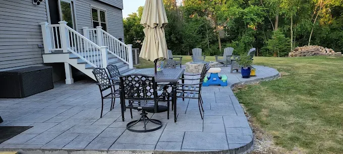 Patio with outdoor dining set and umbrella, next to a house with a staircase and a large yard.