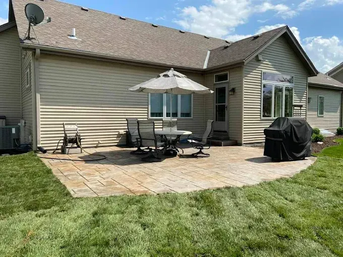 Backyard patio with patio furniture, a grill, and an umbrella, next to a tan house.