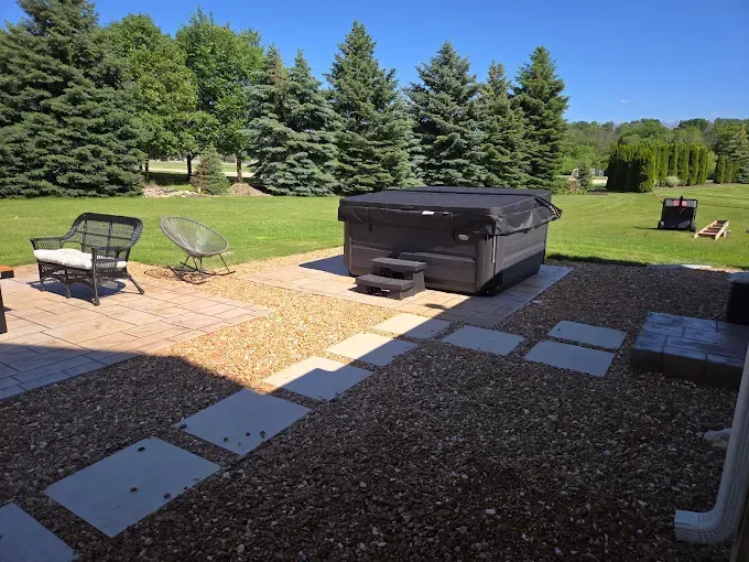 Hot tub on a stone patio with seating, surrounded by gravel and a green lawn with trees.