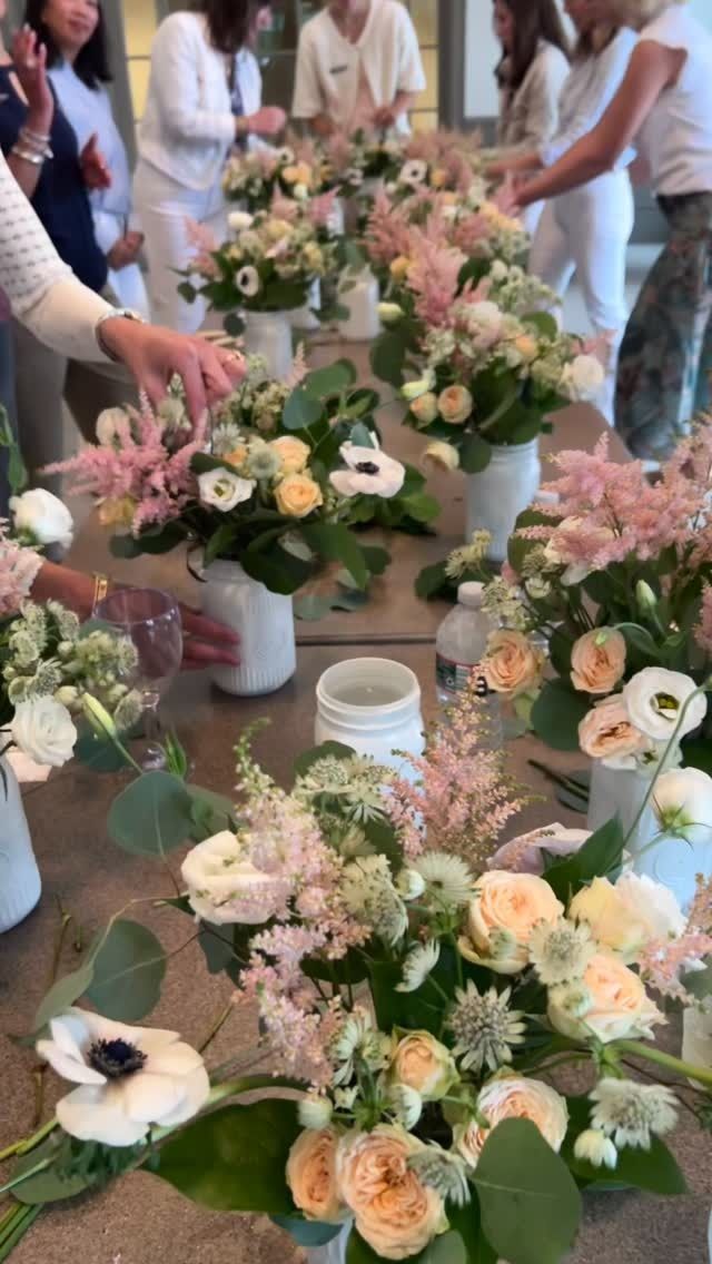 People arranging floral bouquets on a long wooden table. Vases hold arrangements of mixed blooms, including pale pink and white flowers.