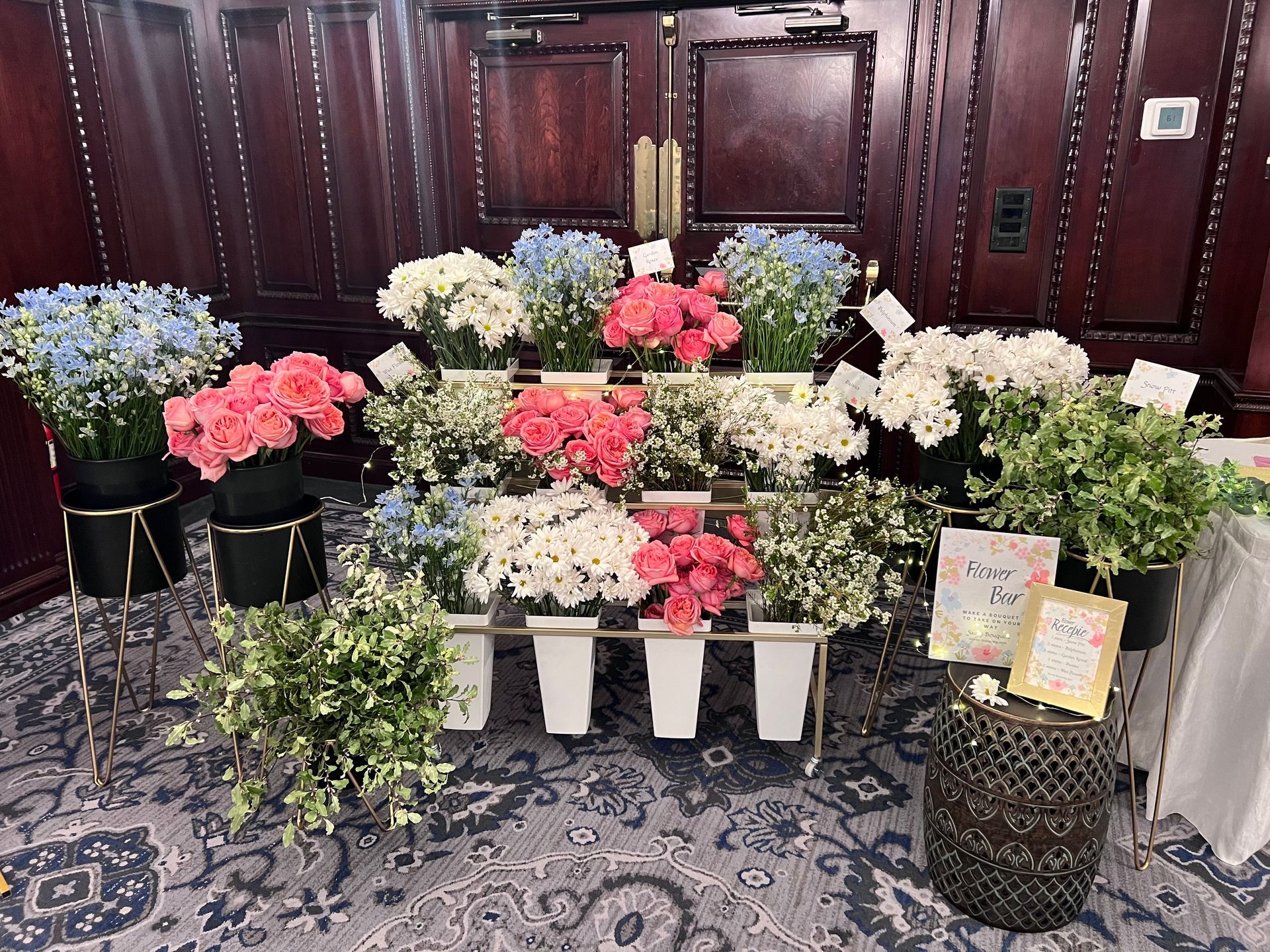 Flower display with various blooms: blue, pink, and white. Gold stands, white buckets. Against wooden wall.