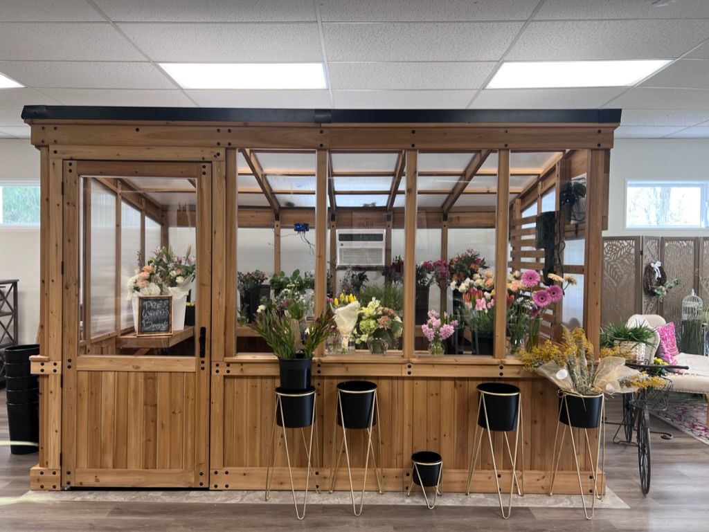 Wooden greenhouse filled with colorful flowers for sale; black pots on stands in front.