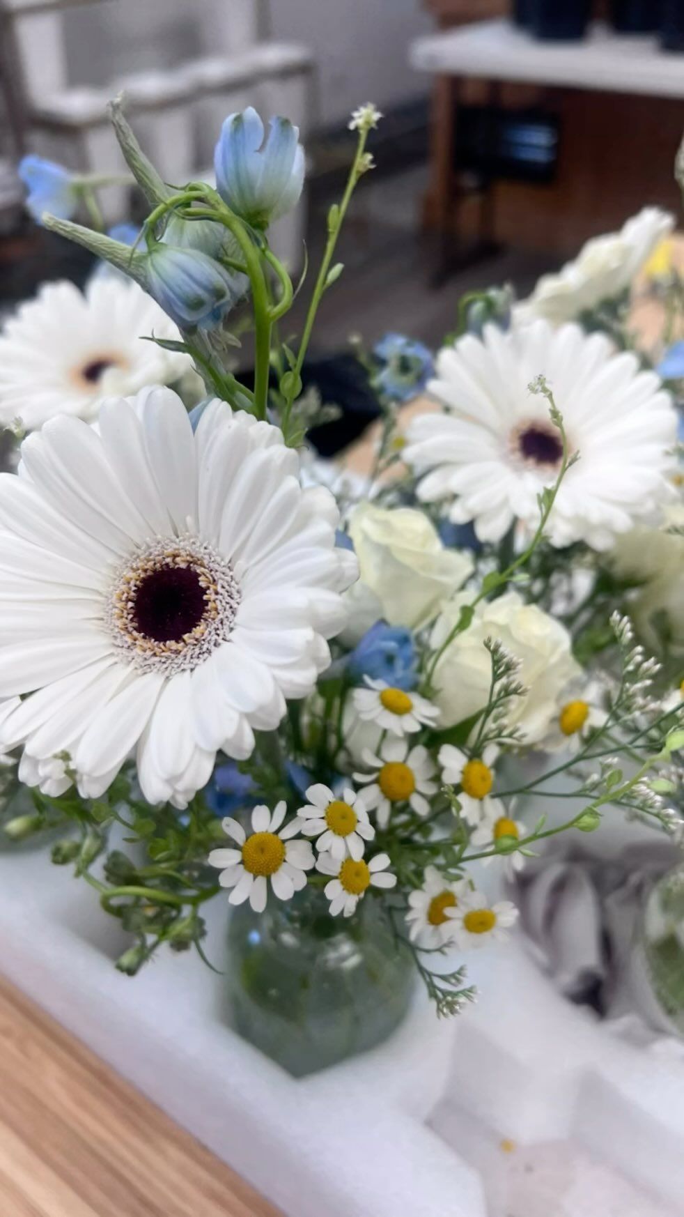 A vase filled with white daisies and blue flowers on a table.
