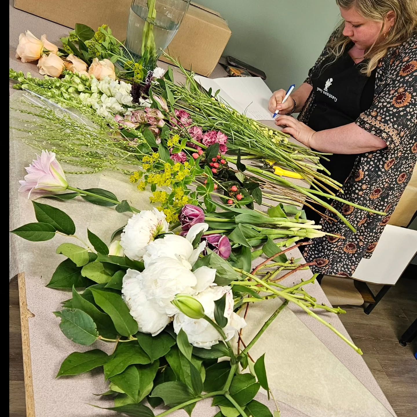 A woman is sitting at a table with flowers and writing on a piece of paper