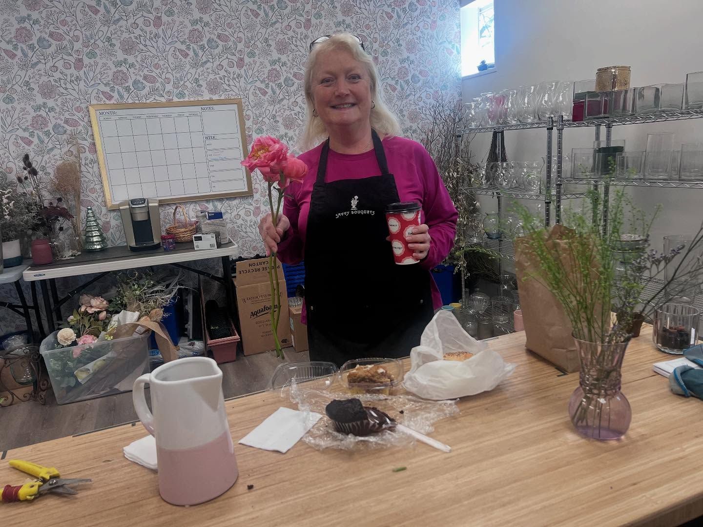 A woman is standing at a table holding a flower and a cup of coffee.