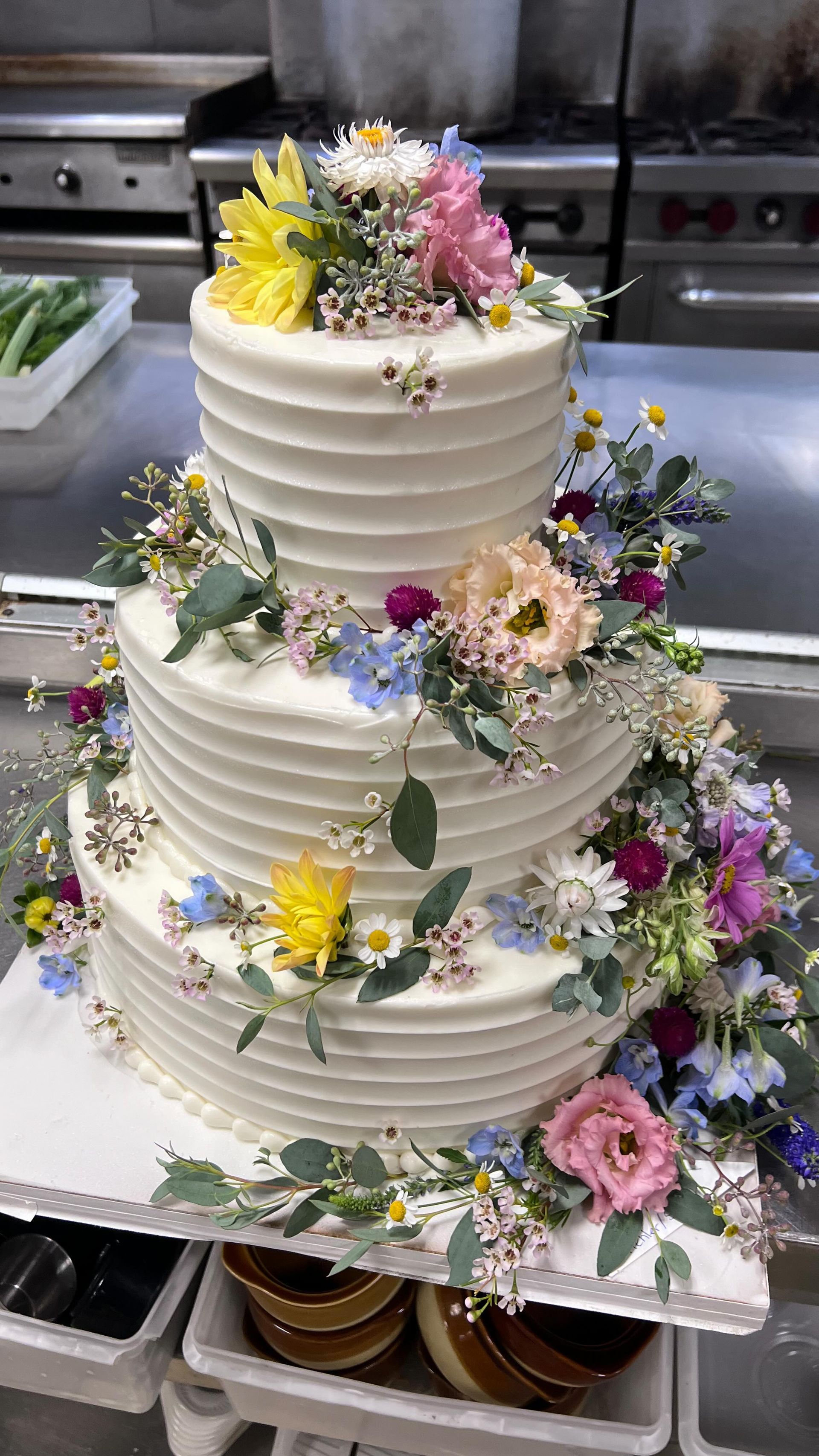 A wedding cake decorated with flowers is sitting on a table in a kitchen.