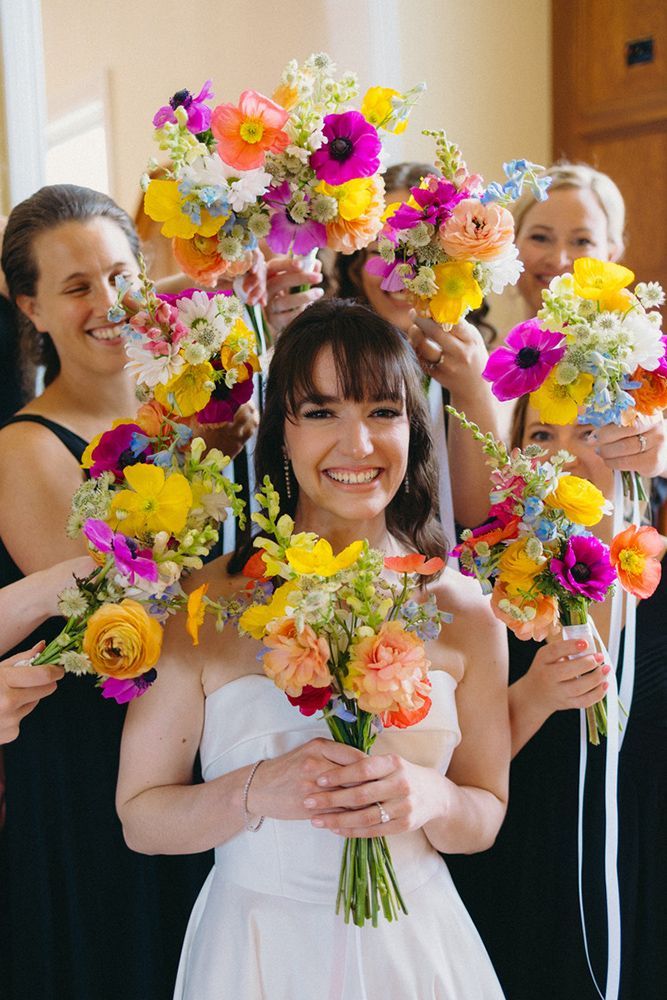 Bride surrounded by bridesmaids holding colorful bouquets. They're smiling, wearing black dresses, and bouquets feature vibrant yellows, oranges, and pinks.