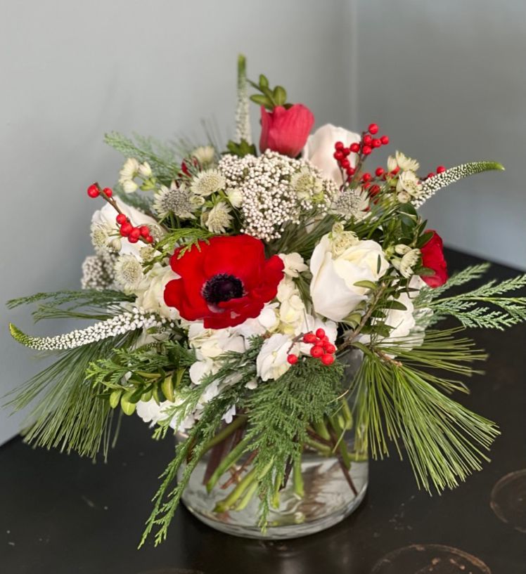 A bouquet of red and white flowers, including anemones and roses, with green foliage in a clear glass vase.