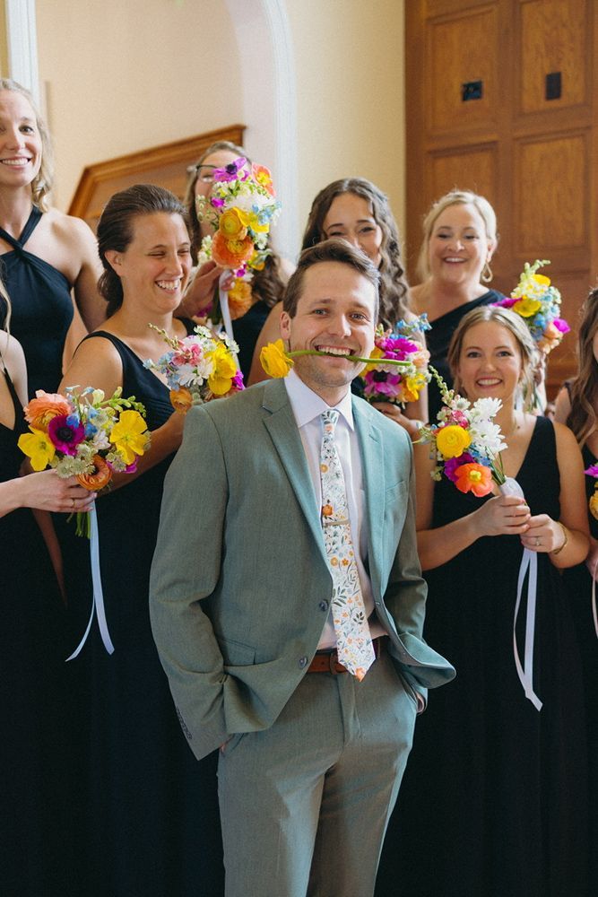 A man in a gray suit with a whimsical tie poses with bridesmaids in black dresses, all holding colorful bouquets indoors.