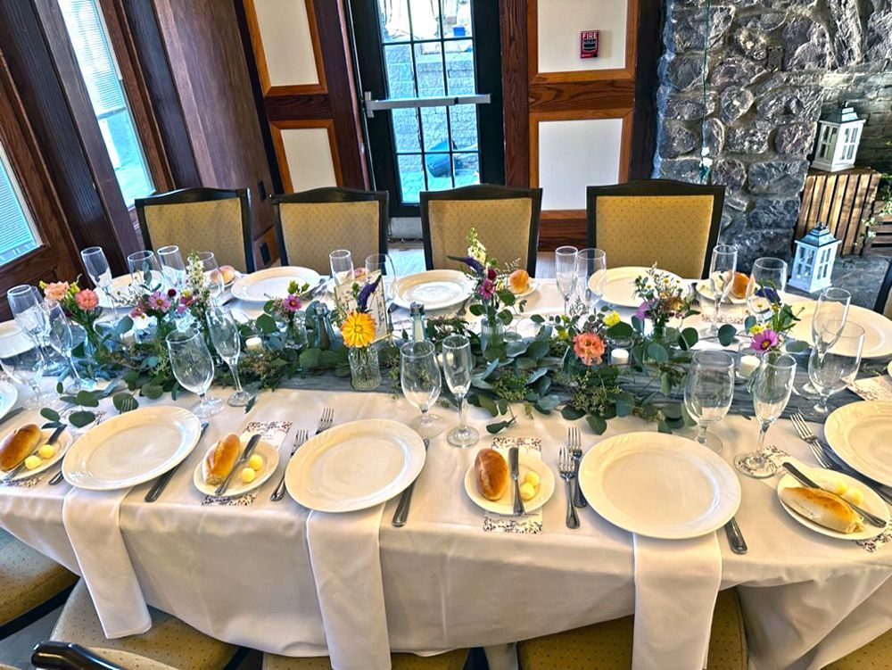 Formal dining table set for a celebration. White tablecloth, floral centerpiece, place settings, bread rolls, and champagne flutes.