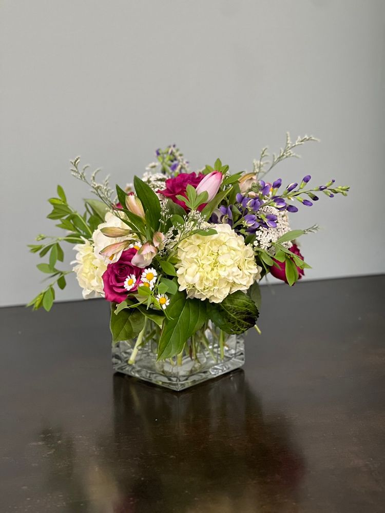 A square glass vase filled with a small floral arrangement featuring cream and purple flowers, surrounded by green foliage, set on a brown surface.