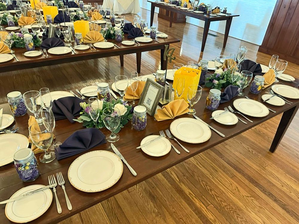 Formal dining tables set for a special event. Brown tables are decorated with white and gold dishes, silverware, and napkins in yellow and dark blue.
