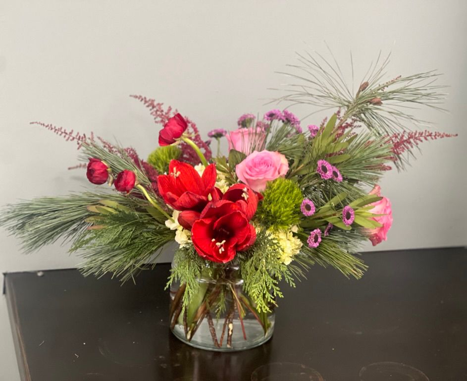 Floral arrangement in a clear vase on a dark surface. It features red, pink, and green flowers with pine branches.