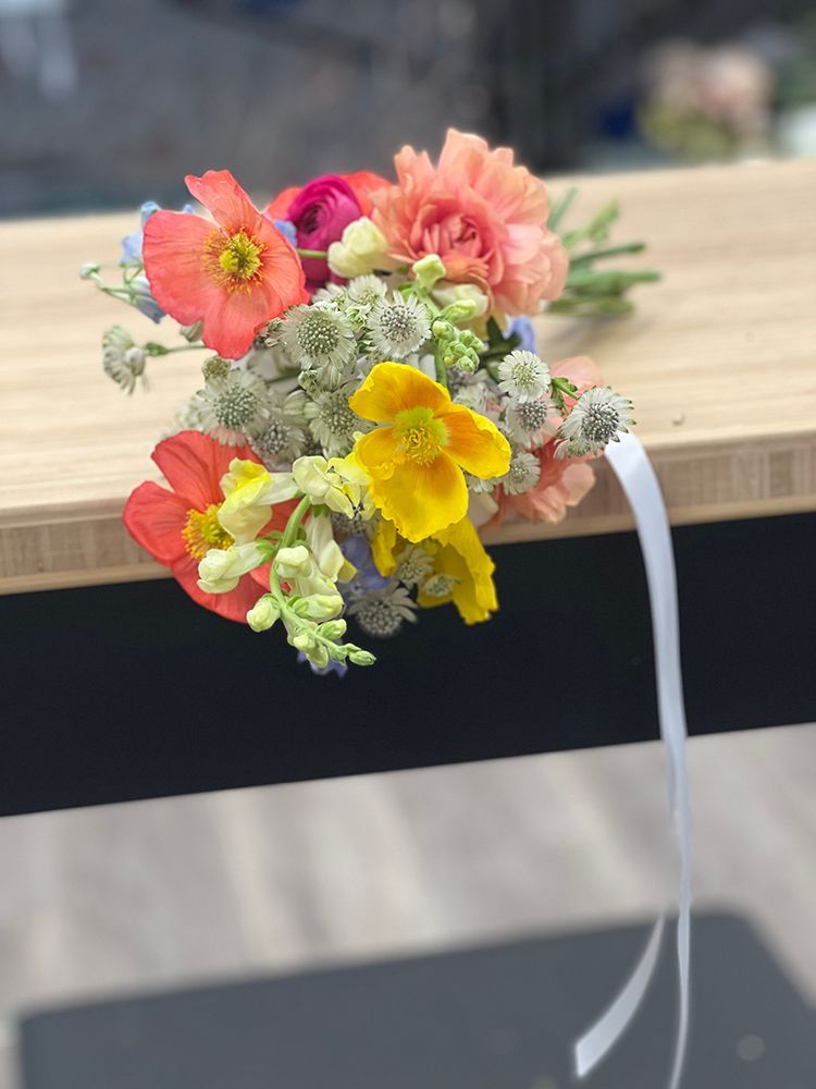 Small bouquet of colorful flowers, including orange poppies and yellow nasturtiums, on a wooden surface with a white ribbon.