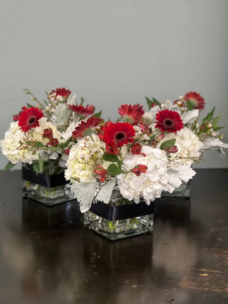 Three floral arrangements in square glass vases with red gerbera daisies and white hydrangeas.