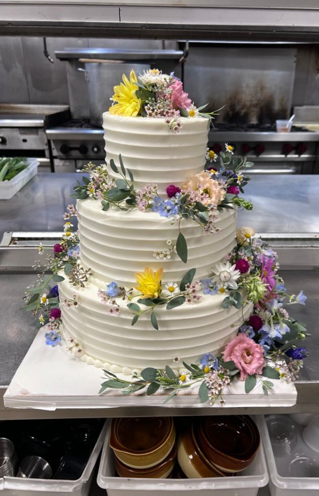 Three-tiered white cake decorated with colorful flowers. Set in a commercial kitchen.