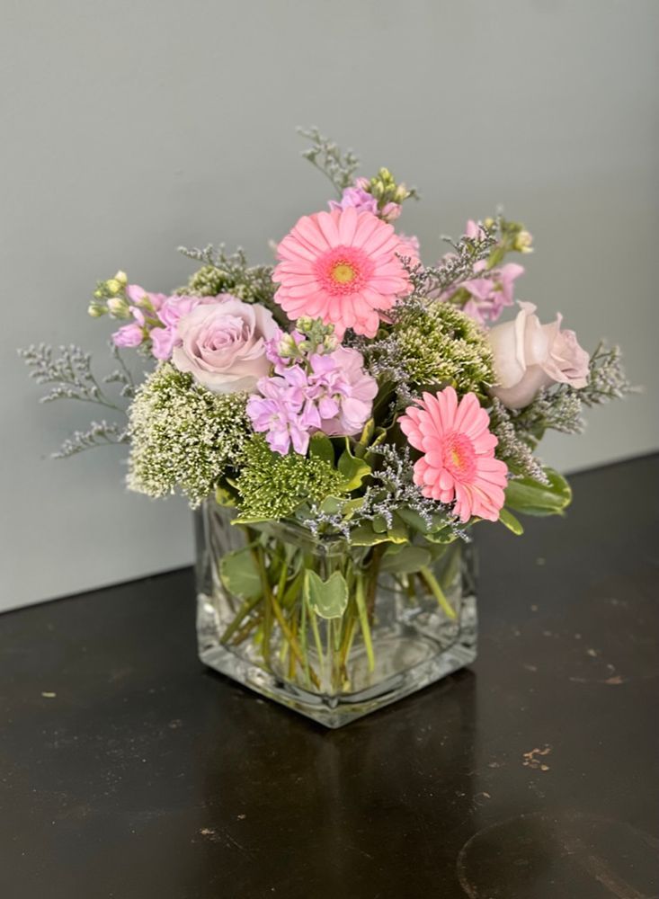 A floral arrangement in a square glass vase features pink gerbera daisies, lavender roses, and greenery.