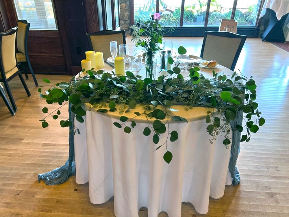Round table with white tablecloth, greenery centerpiece, candles, and chairs in a room with a window backdrop.