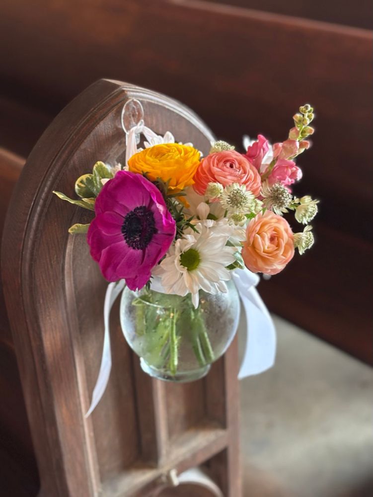 Small floral arrangement in a glass bowl hanging on a wooden church pew, featuring vibrant pink, orange, and white flowers.