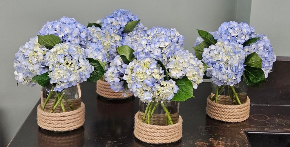 Blue hydrangea flower arrangements in glass jars wrapped with rope, displayed on a dark countertop.