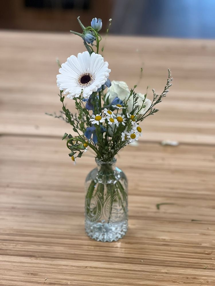 Small flower arrangement in a clear, patterned glass vase on a wooden table; features a white gerbera daisy and small blue and white flowers.
