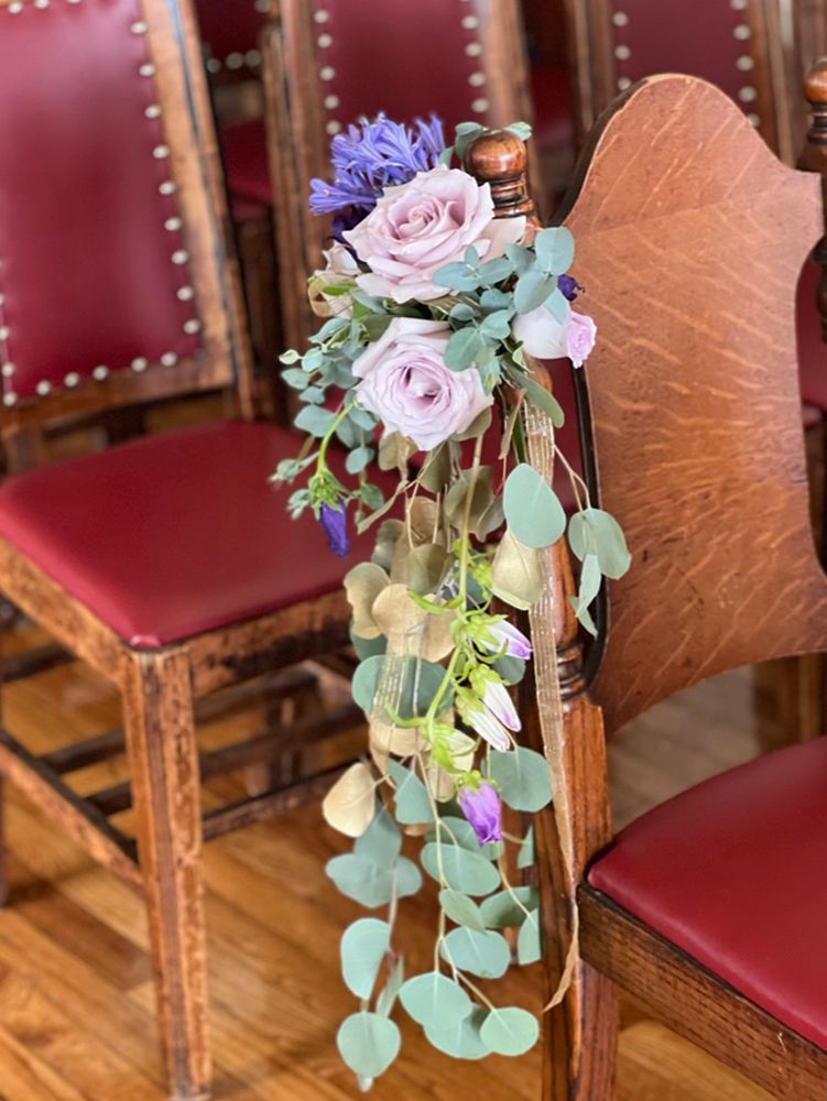 A chair decorated with cascading flowers and greenery, lavender, pink, and green hues. Set in a room with other chairs.