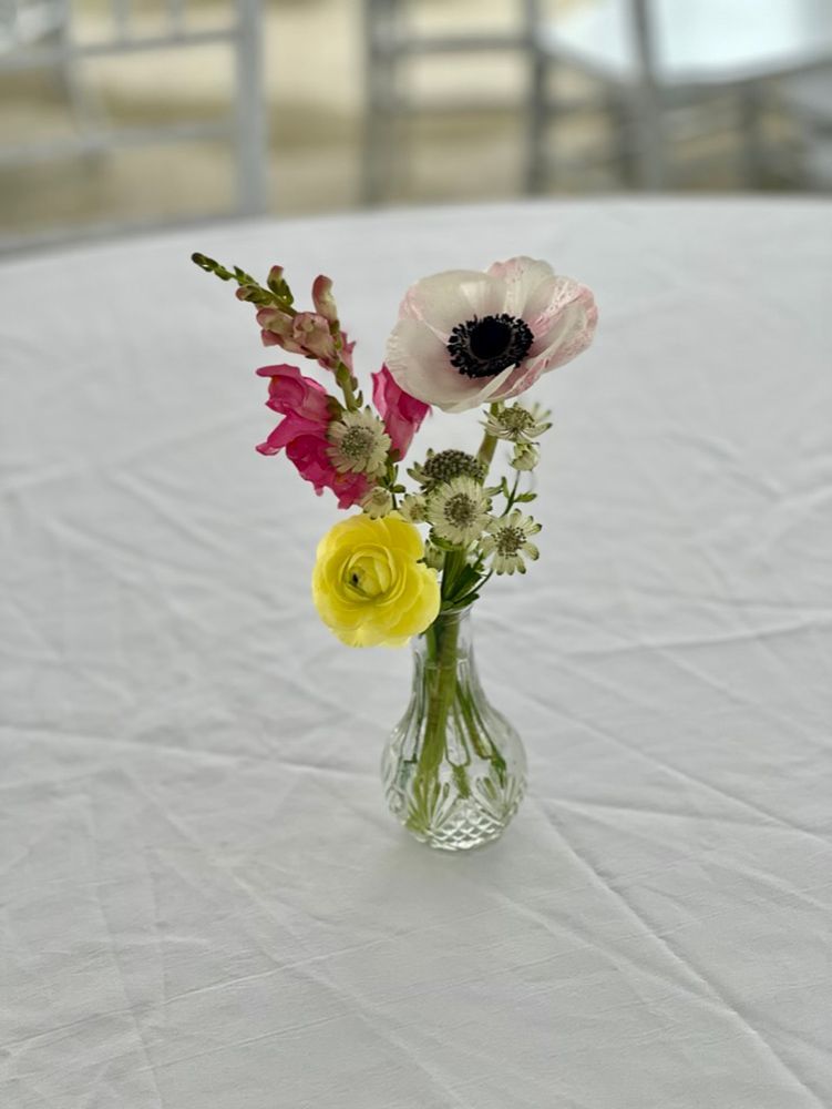 A small bouquet of pink, yellow, and white flowers in a clear glass vase, set on a white tablecloth.