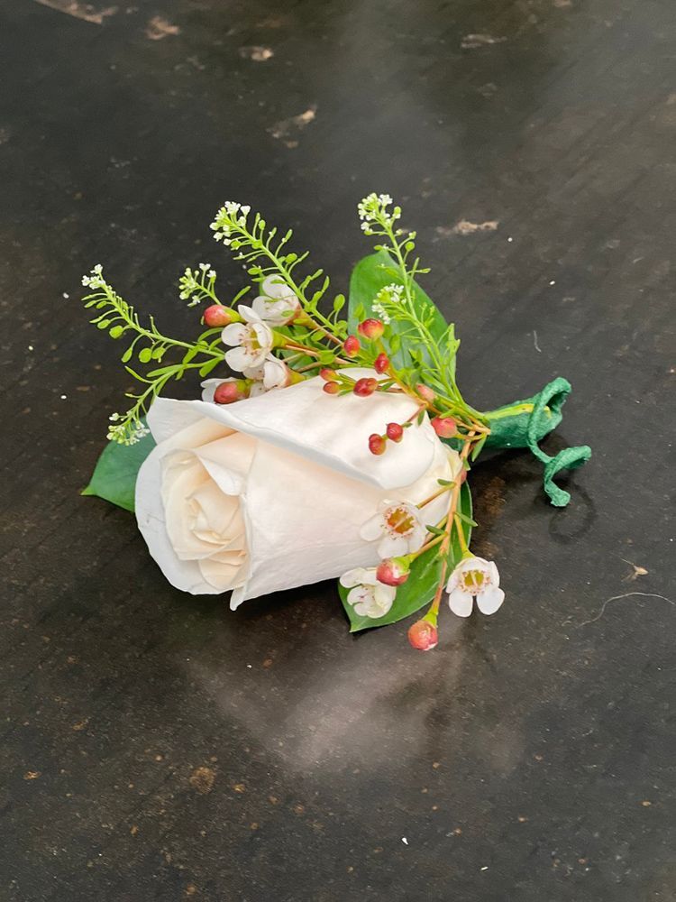 White rose boutonniere with small white flowers, red berries, and green leaves on a dark surface.