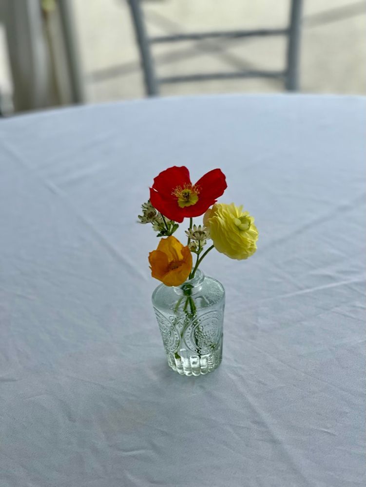 A small glass vase with three colorful flowers (red, yellow, orange) on a white tablecloth.