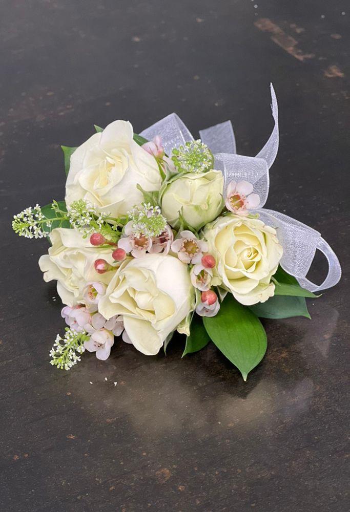 Wrist corsage of white roses and small pink flowers, accented with greenery and a white ribbon on a dark surface.