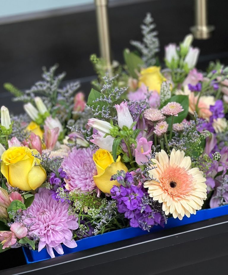 Floral arrangement in a blue rectangular box with yellow roses, purple chrysanthemums, and other colorful flowers.