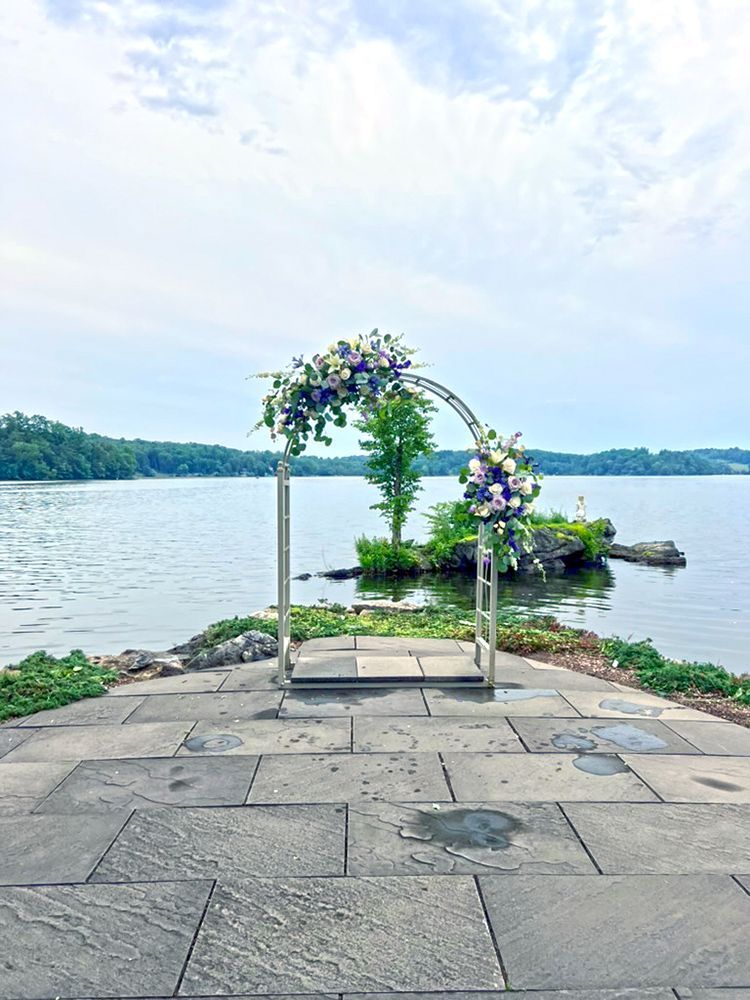 Ceremony arch decorated with flowers, overlooking a lake with an island in the background. The setup is on a stone patio.