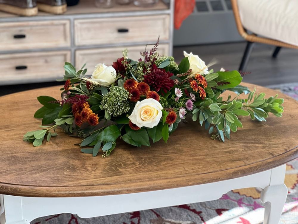 An oval coffee table with a floral arrangement of white roses, dark red mums, and green foliage.