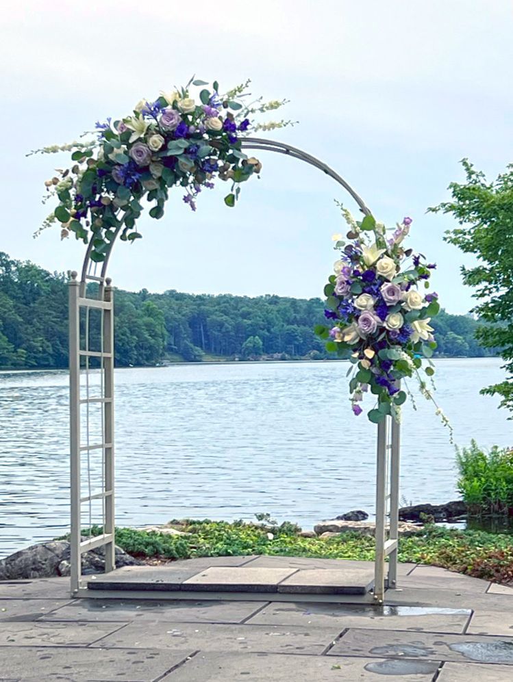 A silver wedding arch decorated with purple and white flowers, set beside a lake with a forest in the background.
