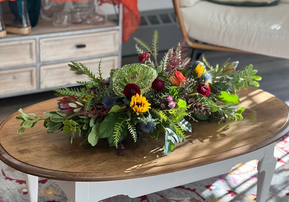 A colorful floral arrangement sits on an oval wooden coffee table in a bright room with a white chair in the background.