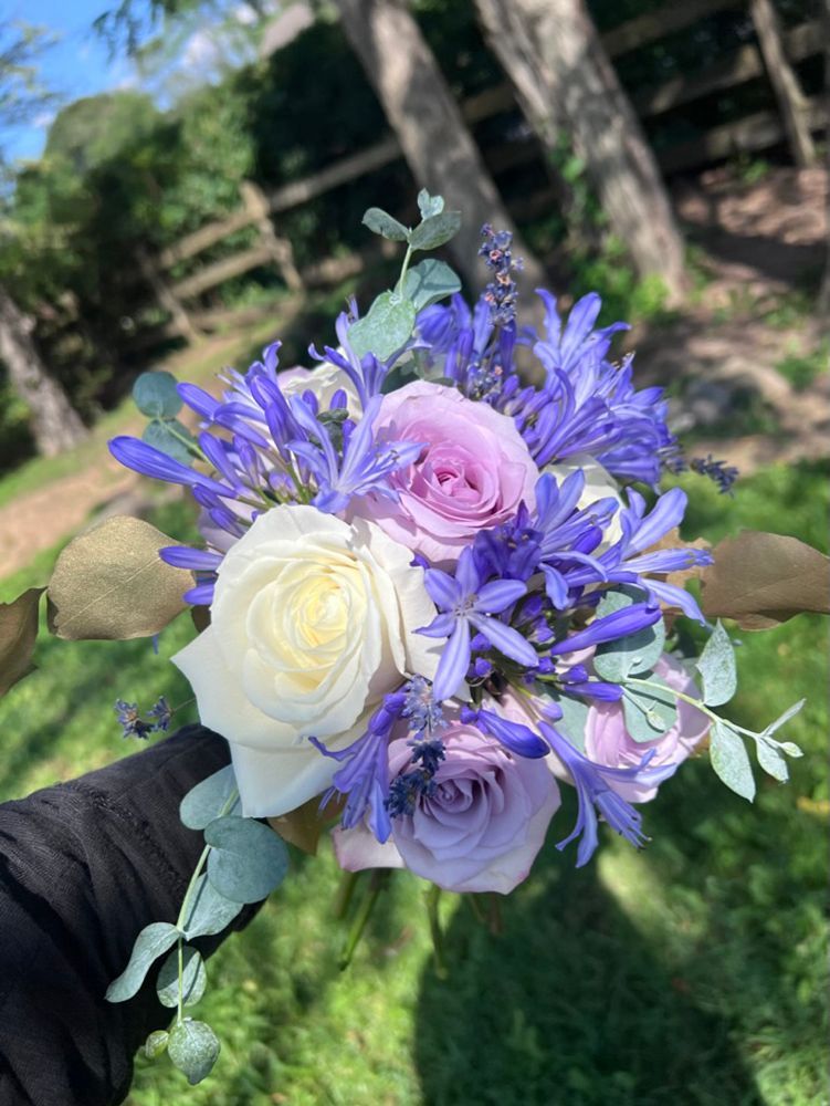 Close-up of a small bouquet with white and purple roses, blue flowers, and green foliage. Held by a person outdoors.