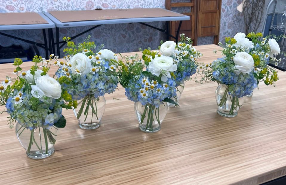 Six floral arrangements in small glass vases sit on a light wood table. The bouquets feature white, blue, and green flowers.