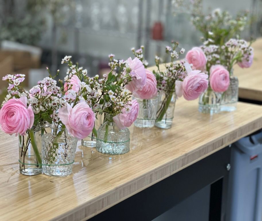 Six small clear glass vases, each with a pink ranunculus and delicate white flowers, lined up on a light wood surface.