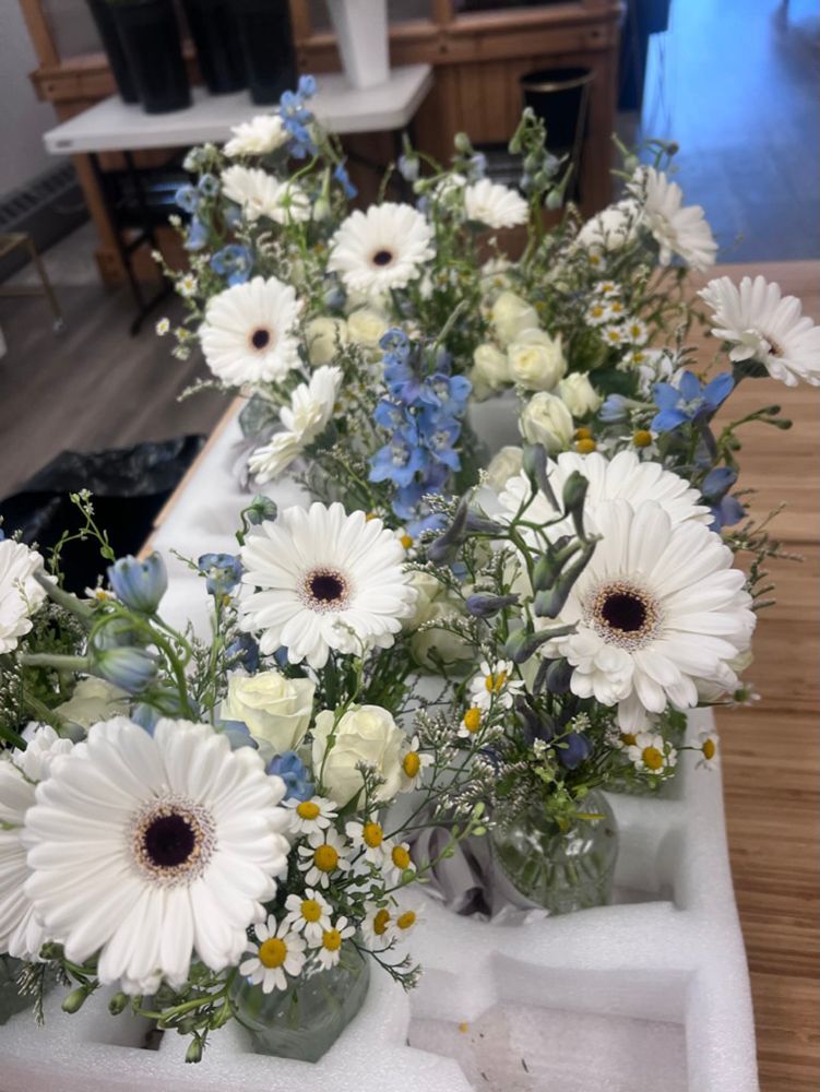 White and blue floral arrangements with daisies and gerbera daisies sit on a table in a shop.