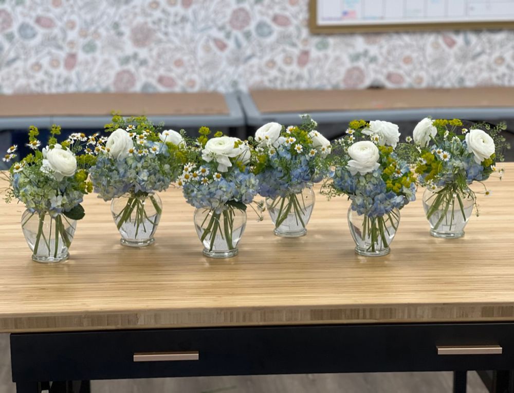 Six floral arrangements with white and blue flowers in glass vases sit on a wooden table.