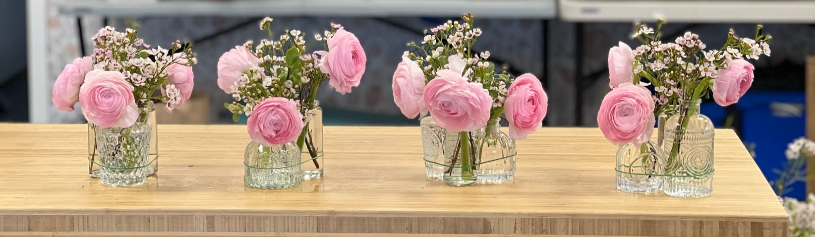Four small glass vases with pink flowers sit on a wooden table.