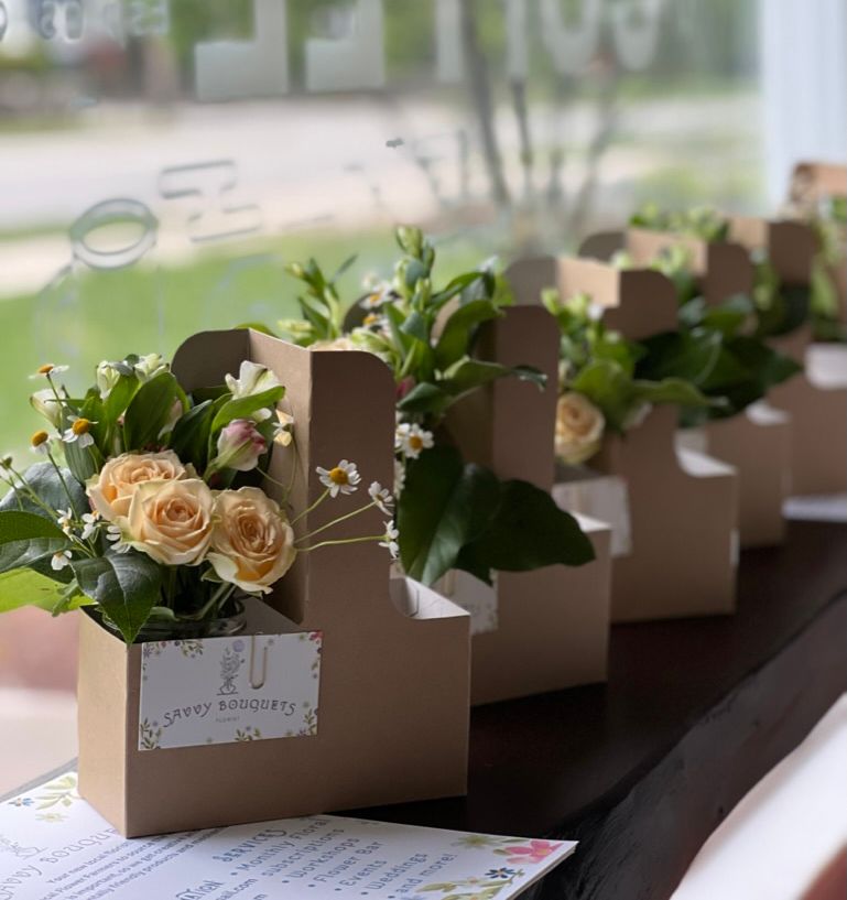 Flower arrangements in cardboard carriers sit on a counter in front of a window. Light yellow roses, greenery, and small white flowers fill the carriers.