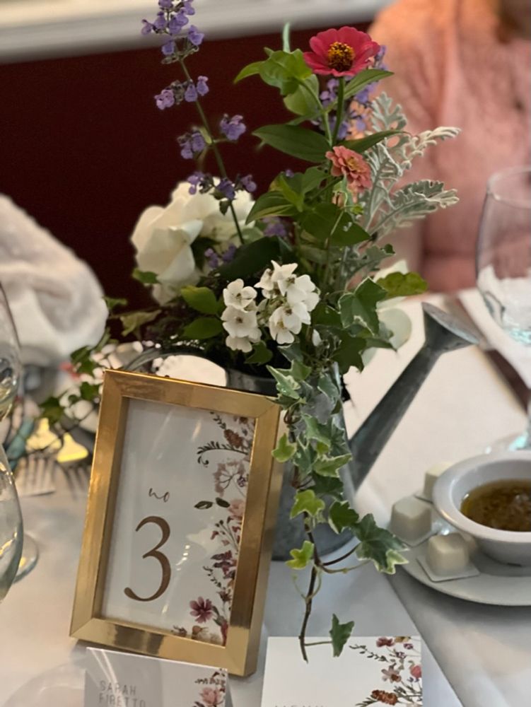 Table setting with floral arrangement in a watering can, table number 3 in a gold frame. Light-colored tablecloth, with a seated person in the background.