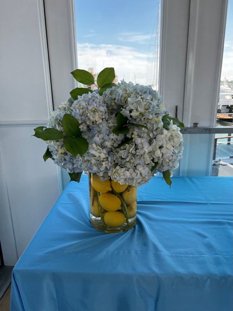 Blue hydrangea flowers in a glass vase filled with lemons on a blue tablecloth; window in background.
