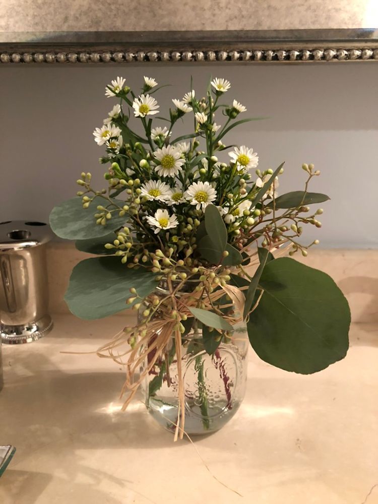 Flower arrangement in a mason jar with white daisy-like flowers and green eucalyptus leaves, tied with a twine bow, sitting on a counter.