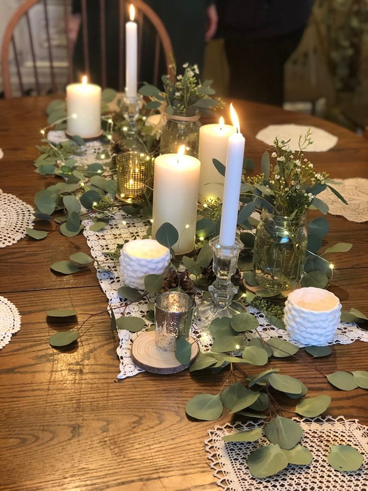 A wooden table centerpiece with lit candles, greenery, and fairy lights. Table is set for a gathering.